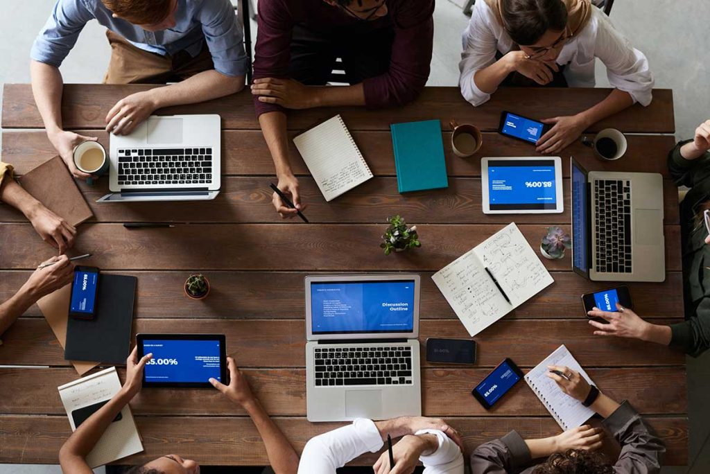 picture from above of eight coworkers sitting together at a table with their laptops, notebooks, and tablets