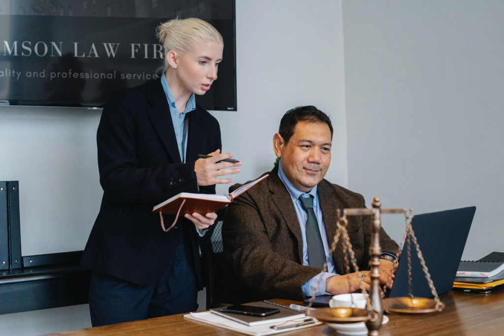A female attorney standing next to a male attorney as they read his laptop.