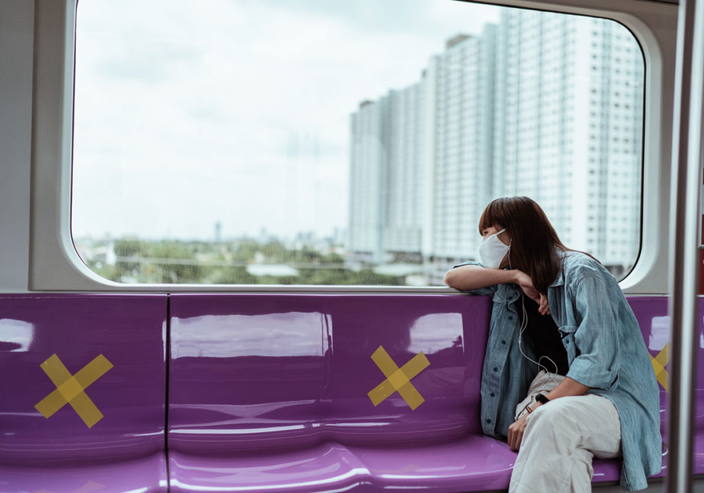 Woman sitting inside a subway car on a purple seat wearing a mask.